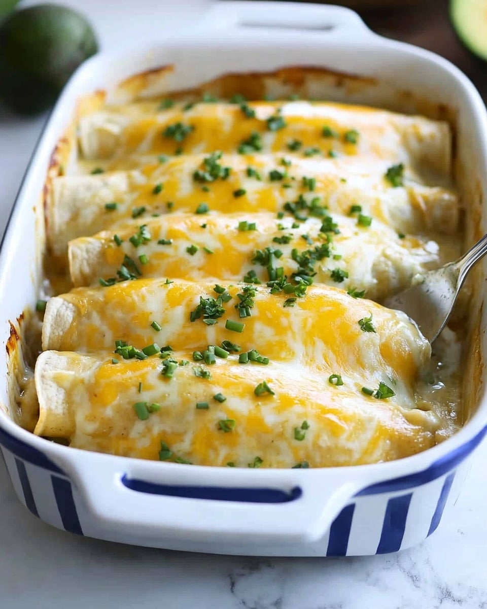 Smothered Green Chile Chicken Burritos, green chile chicken burritos, hearty burrito recipes, easy Mexican chicken burritos, cheesy green chile burritos The dish shows a white baking dish with blue stripes on the outside, filled with five rolled enchiladas covered in melted cheese. Each enchilada has a soft, light yellow tortilla base, covered fully with thick, creamy, white sauce and topped with a golden-yellow melted cheese layer that stretches slightly as one enchilada is lifted by a spatula. Small green chopped chives are sprinkled over the cheese, adding a fresh color contrast. The background is a white marbled surface with blurred avocado halves and a small bowl visible. The