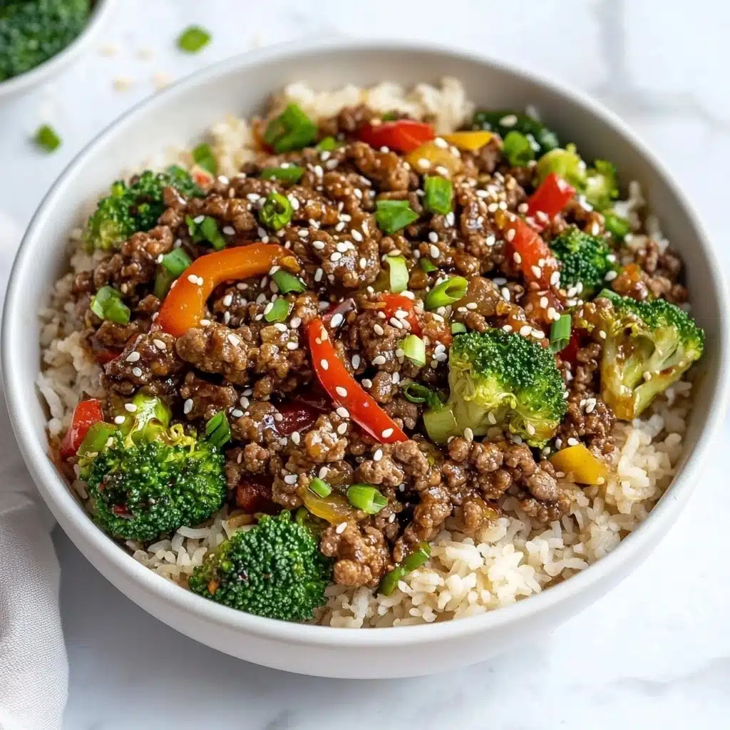Ground Beef and Broccoli Stir Fry, easy ground beef stir fry, quick broccoli stir fry, healthy beef broccoli recipe, Asian-inspired beef stir fry The dish is served in a large white bowl placed on a white marbled surface. The base layer is light brown cooked rice spread evenly around the bowl. On top of the rice, there are pieces of cooked ground beef that appear dark brown and juicy, mixed with bright green broccoli florets and vibrant red bell pepper slices. There are small bits of green onions scattered throughout the dish, adding fresh green specks. White sesame seeds are sprinkled over the beef, adding texture and contrast. The overall look is colorful and fresh, with a mix of soft, tender, and crisp textures.
