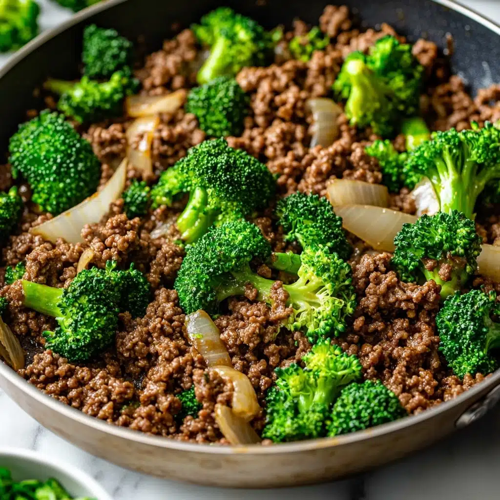 Ground Beef and Broccoli Stir Fry, easy ground beef stir fry, quick broccoli stir fry, healthy beef broccoli recipe, Asian-inspired beef stir fry A close-up view of a pan filled with cooked ground beef that is brown and crumbly as the base layer, mixed with pieces of cooked white onion that are slightly translucent and scattered evenly throughout. On top, bright green broccoli florets with a fresh, crisp texture are mixed in, adding a contrasting color and a fresh look. The pan is placed on a white marbled surface, and a small part of a small bowl with green sliced ingredients is visible near the bottom left corner. The overall scene captures a warm and hearty cooked dish, with a mix of textures from soft beef, tender onions, and fresh broccoli