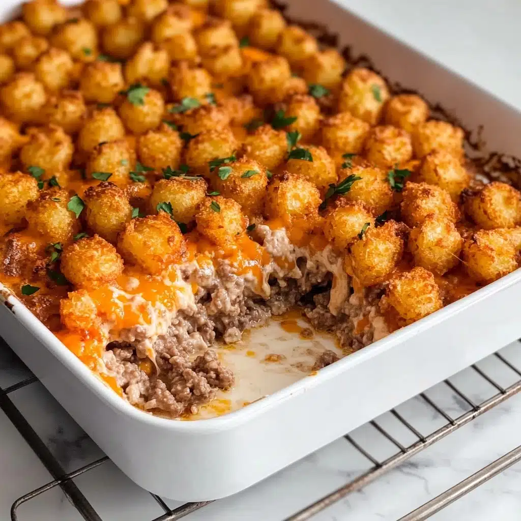 Tater Tot Casserole, easy tater tot casserole, cheesy tater tot bake, hearty dinner ideas, comfort food recipes The image shows a white rectangular baking dish with a layered casserole inside. The top layer is covered with golden-orange roasted tater tots sprinkled with small pieces of green herbs. Beneath the tater tots is a browned layer of ground beef mixed with a creamy white sauce. A portion of the casserole is scooped out from one corner, revealing the two distinct layers. The dish sits on a metal wire rack, all placed on a white marbled surface.