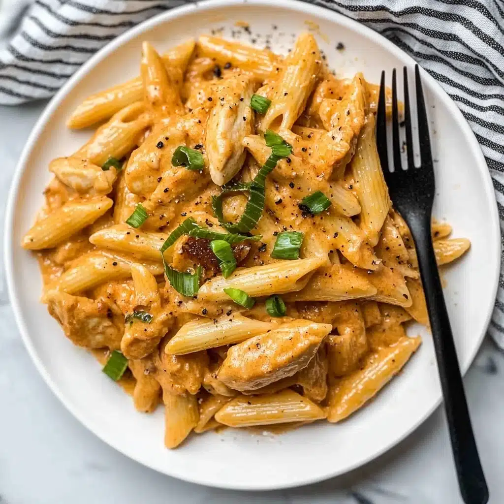 One Pot Creamy Cajun Chicken Pasta, Cajun Chicken Pasta, Easy Cajun Pasta Recipe, Creamy Cajun Pasta, One Pot Pasta Dinner A white round plate holds a creamy pasta dish made of several layers: at the bottom are pieces of light brown cooked chicken and a soft, light orange sauce mixed with whole and chopped penne pasta which is pale yellow. Scattered on top are bright green sliced scallions adding freshness, and small black pepper flakes are sprinkled around. A black fork rests on the right side of the plate touching the pasta, and the whole plate is set on a white marbled surface with a black and white striped cloth nearby.