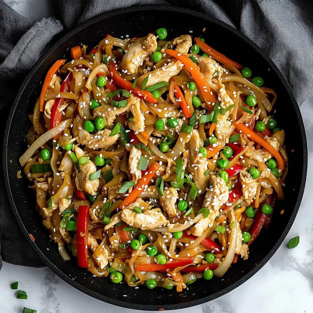 Chicken and Cabbage Stir Fry, healthy chicken stir fry, quick weeknight dinner, easy cabbage stir fry, nutritious vegetable chicken dish This image shows a black pan filled with a colorful stir-fry. The dish has several layers: small chunks of light beige cooked chicken are mixed with thin strips of red bell pepper, orange carrot, and white onion. Bright green peas and chopped green onions are scattered throughout, adding fresh color. White sesame seeds are sprinkled on top, giving a bit of texture and contrast. The pan rests on a white marbled surface with a dark grey cloth nearby.