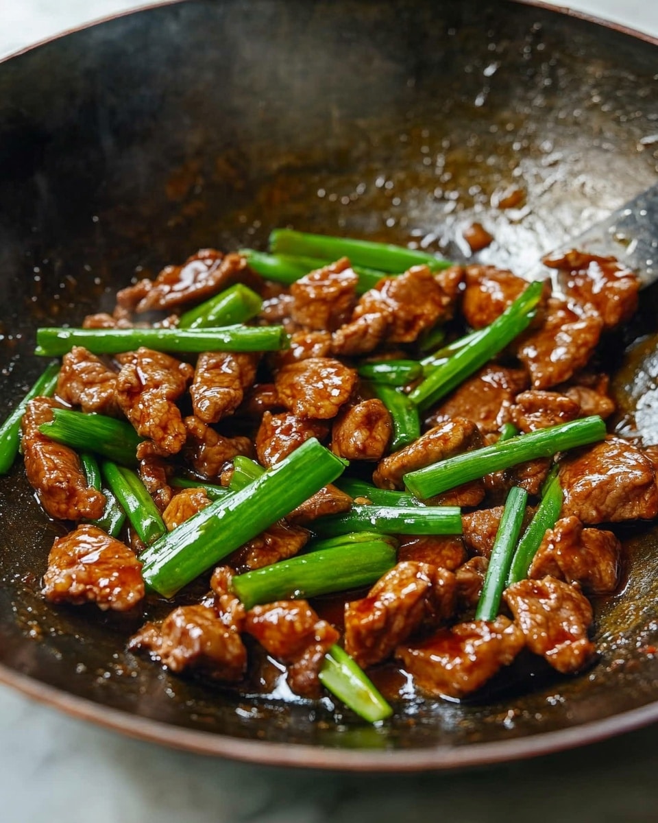 Mongolian Chicken, Chinese stir-fry, crispy chicken recipes, homemade Asian chicken, quick dinner ideas Inside a dark metal wok on a white marbled surface, small pieces of browned meat are mixed with bright green scallions. The meat looks shiny and cooked in a thick brown sauce that pools slightly at the bottom. The scallions are cut into long thin pieces and scattered evenly on top, adding a fresh contrast in color and texture. The wok shows signs of use, with some light marks and a textured surface inside, and a metal spatula edge is visible at the top right corner.