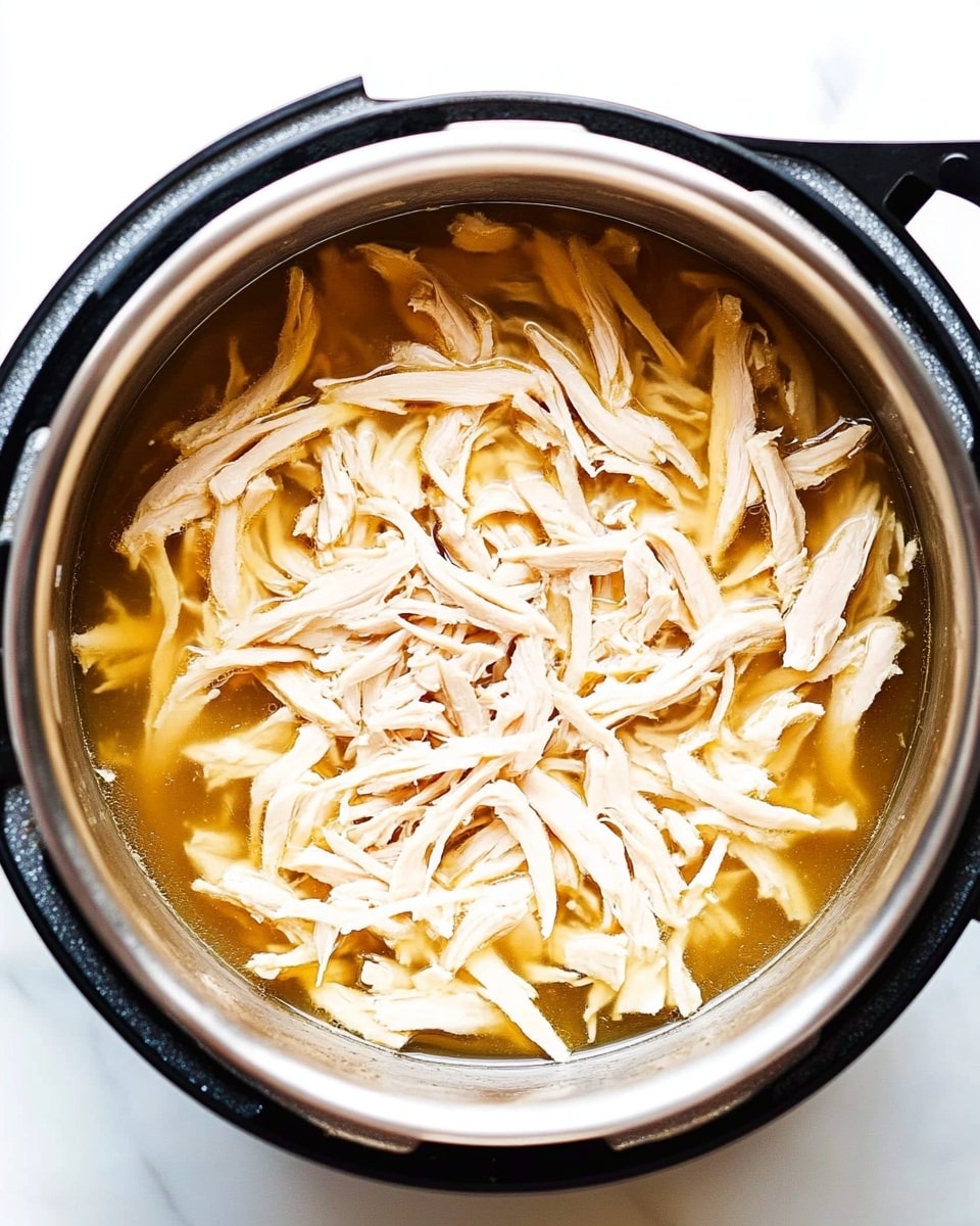 Instant Pot Shredded Chicken, easy shredded chicken recipe, quick chicken dinner, tender shredded chicken, healthy Instant Pot chicken The image shows a close-up top view of shredded light beige chicken resting in a shallow layer of golden-brown broth inside a shiny silver metal pot. The chicken pieces are long, thin, and stringy, scattered unevenly throughout the pot. The pot has a black outer rim and handles, and it sits on a bright white marbled surface. The light reflects softly off the broth and the moist chicken, creating a fresh and clean look.