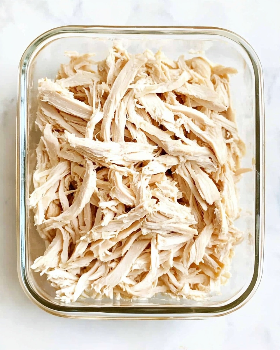 Instant Pot Shredded Chicken, easy shredded chicken recipe, quick chicken dinner, tender shredded chicken, healthy Instant Pot chicken The image shows a clear glass rectangular container filled with shredded cooked chicken. The shredded chicken is light beige, with soft, fibrous texture pieces layered loosely to fill the container evenly from top to bottom. The chicken strands are thin and varied in size and shape, creating a natural, slightly uneven surface. The container sits on a white marbled surface.