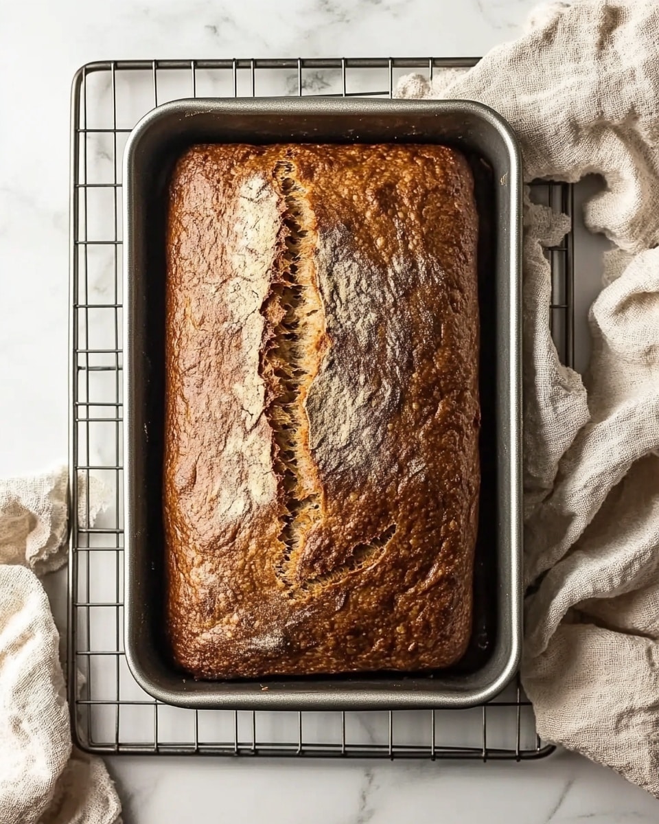 Fast & Moist Banana Bread, quick banana bread, easy banana bread recipe, moist banana loaf, homemade banana bread A rectangular loaf of brown bread with a cracked top surface rests in a silver metal loaf pan, placed on a metal cooling rack over a white marbled texture. The bread's crust is evenly baked with a golden brown color and some darker spots, showing a rough texture. Next to the pan, a beige fabric cloth is casually placed, adding a soft touch to the scene.