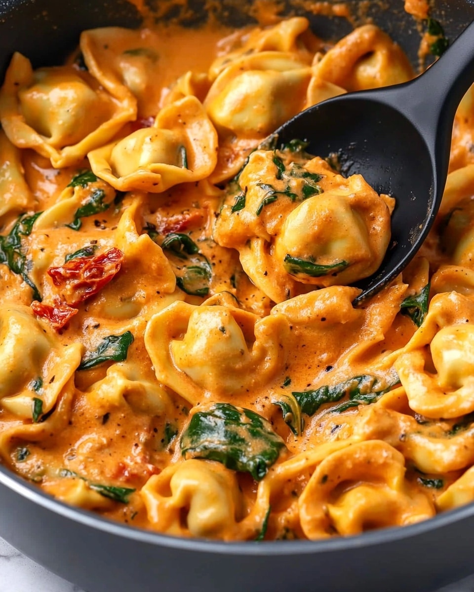 A close-up view of tortellini pasta coated in a rich, creamy orange sauce with visible pieces of green spinach leaves mixed throughout. The tortellini are plump and soft with curved edges, layered naturally inside a gray pan. The sauce has a thick, smooth texture with small bits of red sun-dried tomato and herbs scattered throughout. A black spoon is partially submerged on the right side, lifting some pasta, with the creamy sauce clinging to it. The background features a white marbled texture. photo taken with an iphone --ar 4:5 --v 6.1