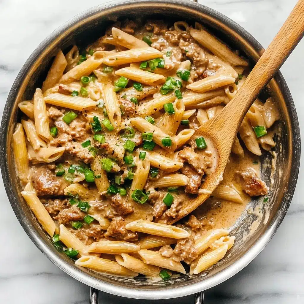 One Pot Creamy Cajun Chicken Pasta, Cajun Chicken Pasta, Easy Cajun Pasta Recipe, Creamy Cajun Pasta, One Pot Pasta Dinner A stainless steel pan filled with cooked penne pasta mixed with small pieces of browned cooked meat and chopped green onions scattered on top. The pasta and meat are coated in a creamy light brown sauce that looks smooth and rich. A wooden spoon rests inside the pan on the right side, partially covered with the pasta mixture. The pan is placed on a white marbled surface.