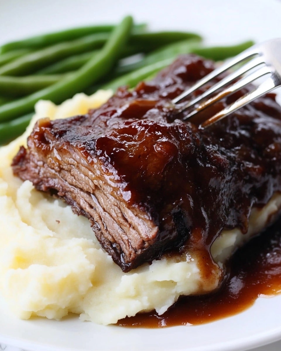 Instant Pot Beef Ribs, Instant Pot Beef Ribs recipe, quick beef ribs, tender beef ribs, easy pressure cooker beef ribs The image shows a white plate with three food layers and a utensil in front. The bottom layer is mashed potatoes, which look smooth and creamy with a soft, fluffy texture, colored white to light yellow. On top of the mashed potatoes is a thick, dark brown barbecued meat piece with a shiny, caramelized sauce glaze and a slightly rough texture. To the right of the plate, green beans add a fresh and crisp green layer with a smooth surface. The background features a white marbled table surface and a blurred kitchen appliance, creating a cozy kitchen feel.