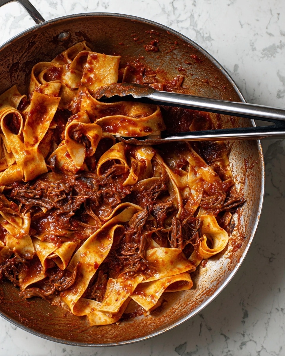 The image shows a large pan with thick, flat pasta ribbons mixed with a rich, dark red tomato sauce. The sauce has chunky bits of dark brown shredded meat and small pieces of tomato, coating the pasta unevenly. Two metal tongs with black rubber tips rest inside the pan, partly covered in sauce. The background is a white marbled surface, giving a clean and bright contrast to the deep colors of the dish. photo taken with an iphone --ar 4:5 --v 7
