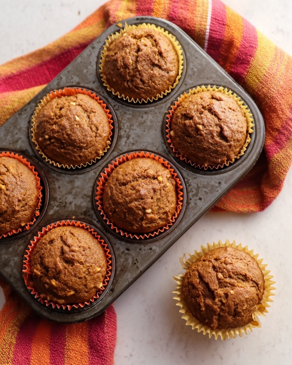 This image shows several pumpkin nut muffins, each with a rough, cracked top and a deep orange-brown color. Six muffins are still inside a dark metal muffin tray lined with orange and yellow paper cups, which have a slightly ruffled edge. Two muffins are outside the tray on a white marbled surface, one still in its peeled-back paper cup and one without the paper. Near the muffins, there is a soft cloth with broad stripes in warm colors like red, orange, and yellow, adding a cozy touch to the scene. The setup gives a warm, fresh-baked feeling. Photo taken with an iphone --ar 4:5 --v 7