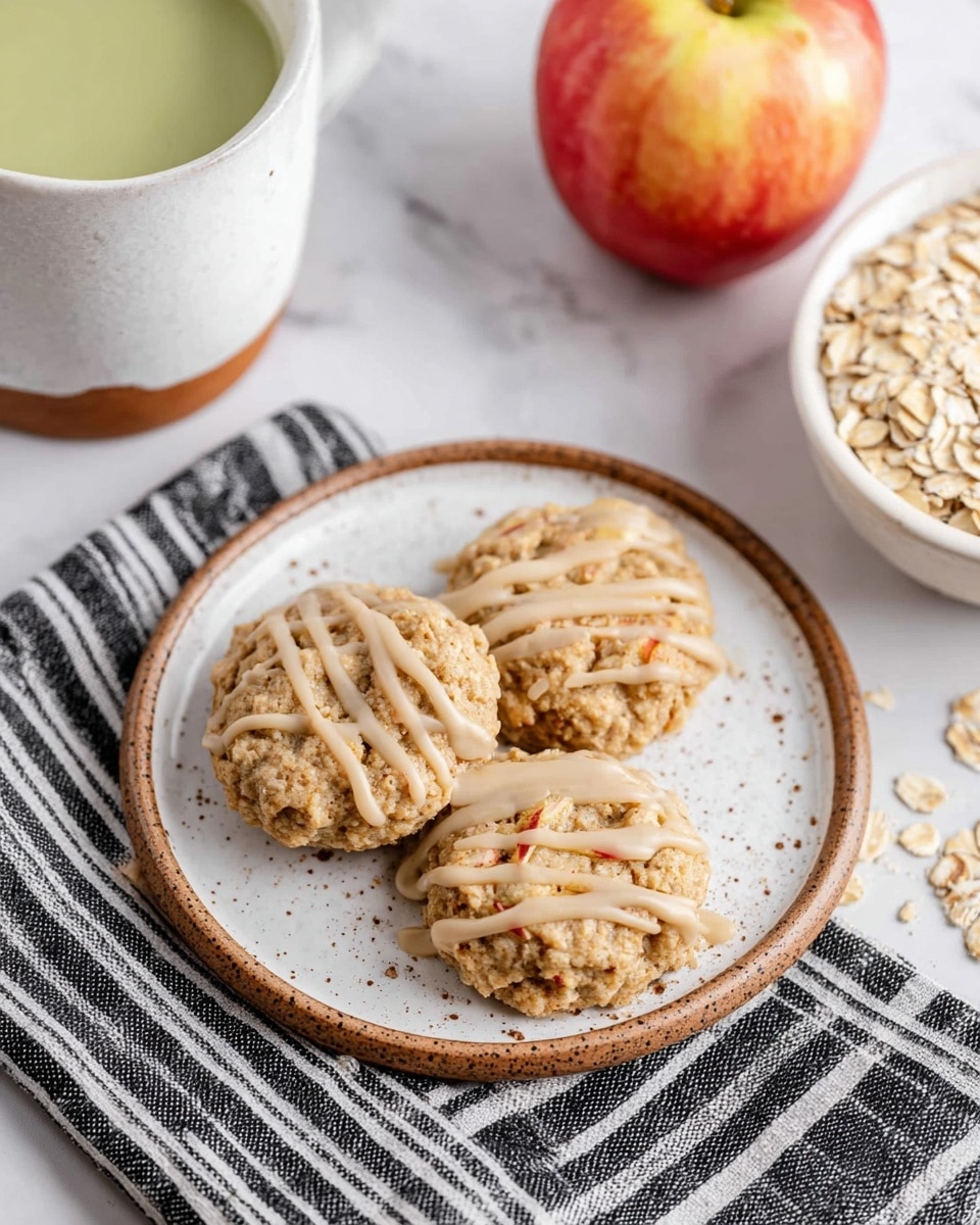 A white speckled round plate filled with about eight rough-textured oatmeal cookies that have small pieces of red apple visible inside; each cookie is drizzled with light beige icing in thin streaks across the top, and the plate sits on a wooden board placed on a white marbled surface; in the background, there is a white cup containing a light green beverage, and a part of another white plate with a cookie is visible, all softly lit by natural light, photo taken with an iphone --ar 4:5 --v 7