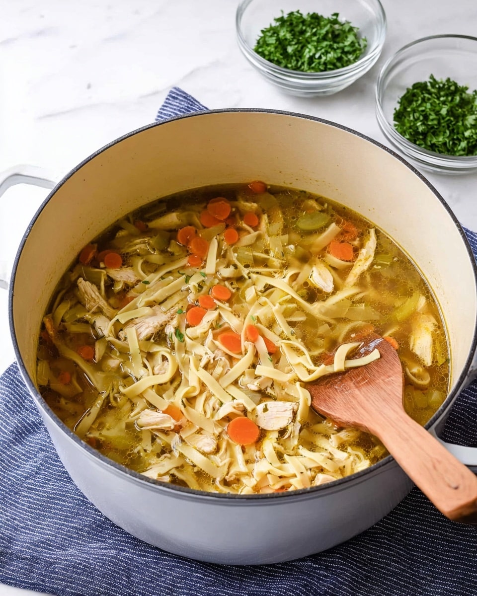 A large white pot filled with clear yellow broth layered with light yellow flat noodles, small orange carrot slices, and bits of light brown chicken, along with small green herb pieces scattered throughout. A wooden spoon with a smooth brown handle rests inside the pot, partially submerged in the soup. In the background, there is a white marbled surface with fresh green parsley and two white bowls stacked on top of each other, with a piece of light brown bread partially visible. Photo taken with an iphone --ar 4:5 --v 7