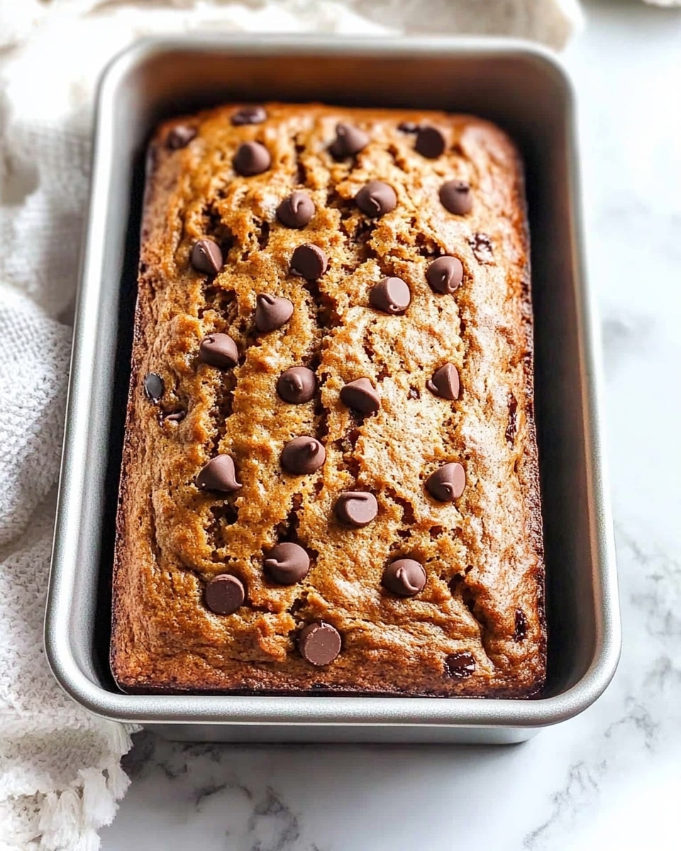 A rectangular silver metal loaf pan filled with a golden brown baked banana bread topped with scattered dark brown chocolate chips, the top of the bread has a textured, slightly cracked surface showing a soft, moist interior. The pan is placed on a white marbled surface, next to a white cloth with small pom-pom details. photo taken with an iphone --ar 4:5 --v 6.1