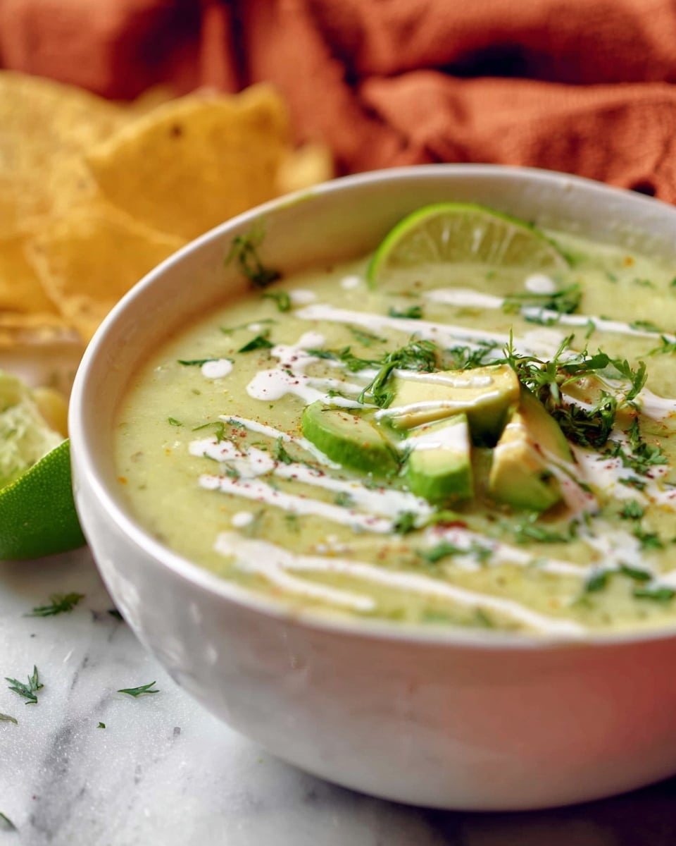 A close-up of a white bowl filled with a creamy light green soup that has a smooth texture with small chunks visible. The soup is topped with sliced avocado pieces, chopped green herbs, and thin white cream drizzles. A green lime wedge rests on the edge of the bowl. In the background, there are a few yellow tortilla chips leaning on the bowl, and a soft orange cloth is also visible. The bowl is placed on a white marbled surface. Photo taken with an iphone --ar 4:5 --v 7