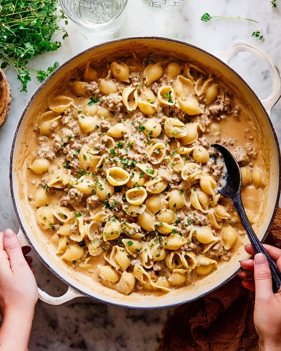 A large white pan filled with three layers: the bottom layer is a thick creamy sauce in light brown, the middle layer consists of small shell pasta pieces mixed with tiny bits of ground meat in light beige with spots of darker brown, and the top layer is lightly sprinkled with small green herb leaves. A woman's hand is holding the pan handle on the bottom left, and another woman's hand is holding a black spoon into the pasta on the top right. The pan sits on a white marbled surface with some green herb sprigs and a clear glass of water nearby. Photo taken with an iphone --ar 4:5 --v 7