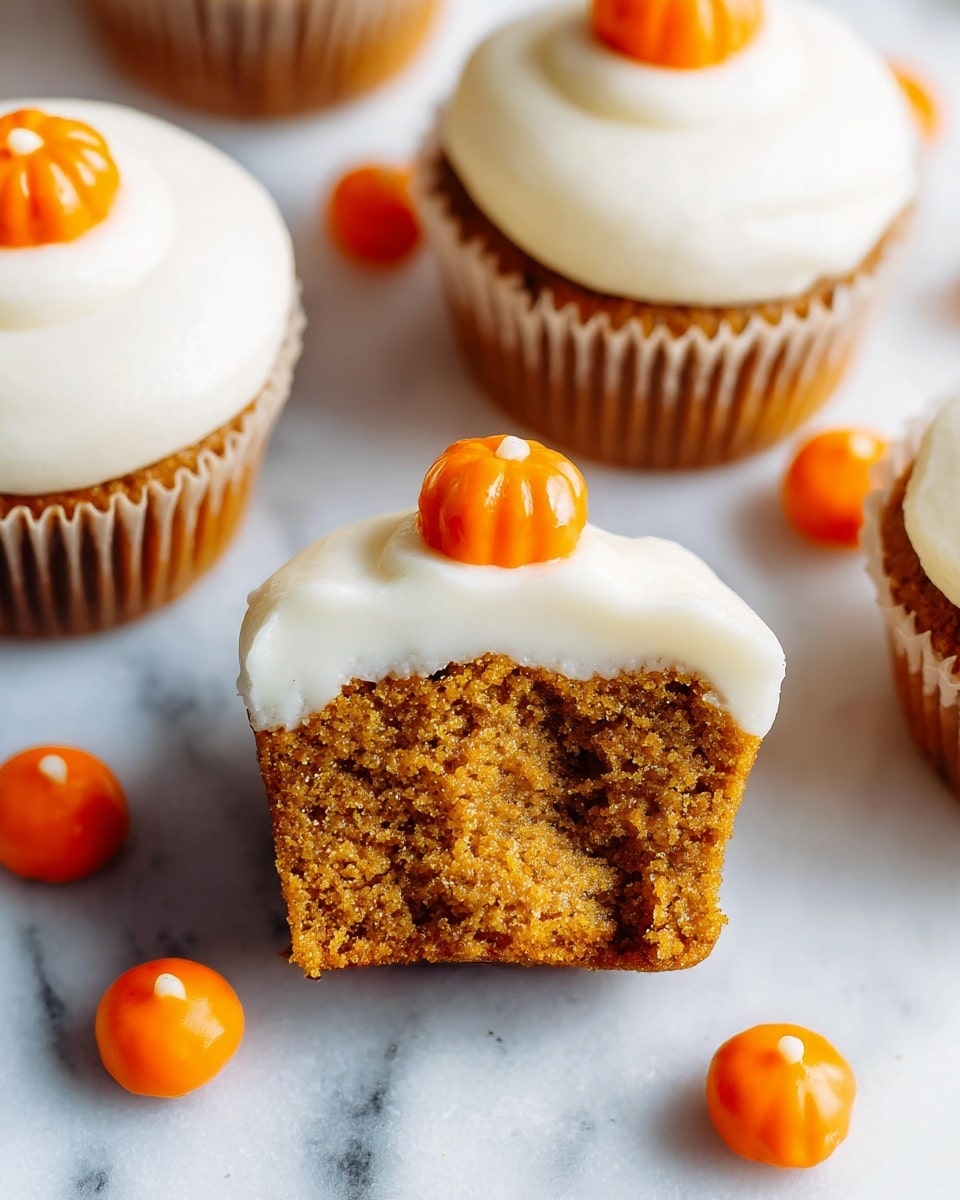 The image shows pumpkin cupcakes with three layers: the bottom layer is a moist, brown-orange pumpkin cake, the middle layer is a thick, smooth white cream cheese frosting, and the top layer is a small, bright orange pumpkin candy decoration. One cupcake is shown with a bite taken, revealing the soft texture inside. The cupcakes are arranged on a white marbled surface, with several loose orange pumpkin candies scattered around. The photo is bright and focused on the cupcakes, showing their texture clearly. photo taken with an iphone --ar 4:5 --v 7