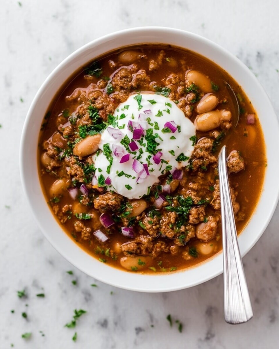 A white bowl filled with a rich brown chili soup that has white beans and small pieces of browned ground meat spread throughout the broth. On top, there is a dollop of white sour cream placed in the center, sprinkled with small pieces of bright green chopped herbs and finely chopped purple onions scattered around. A silver spoon rests inside the bowl, partly submerged in the soup, with its handle leaning out. The bowl is set on a white marbled surface with tiny bits of green herbs scattered around. photo taken with an iphone --ar 4:5 --v 7
