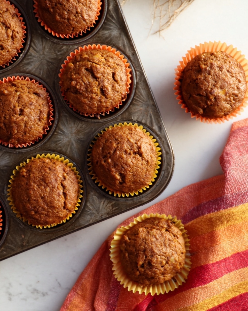 The image shows several brown muffins with a slightly rough, crumbly top texture. The front muffin is partially wrapped in an orange muffin liner with yellow heart shapes, which is peeled back around the base. The other muffins are fully wrapped in the same liners and are placed in a white muffin tray in the background. The surface underneath is a white marbled texture, and there is a folded orange and yellow striped cloth to the right of the muffins. The lighting is natural and soft. photo taken with an iphone --ar 4:5 --v 7