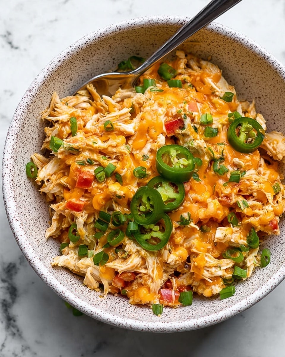 A white bowl filled with shredded white chicken mixed with small pieces of red and orange bell peppers and cooked onions, covered with melted orange cheddar cheese dripping over the top. Small slices of green jalapeño and chopped green onions are scattered on the dish. A silver spoon is placed inside the bowl, resting against the side. The bowl sits on a white marbled surface with green onion stalks blurred in the background. Photo taken with an iphone --ar 4:5 --v 7