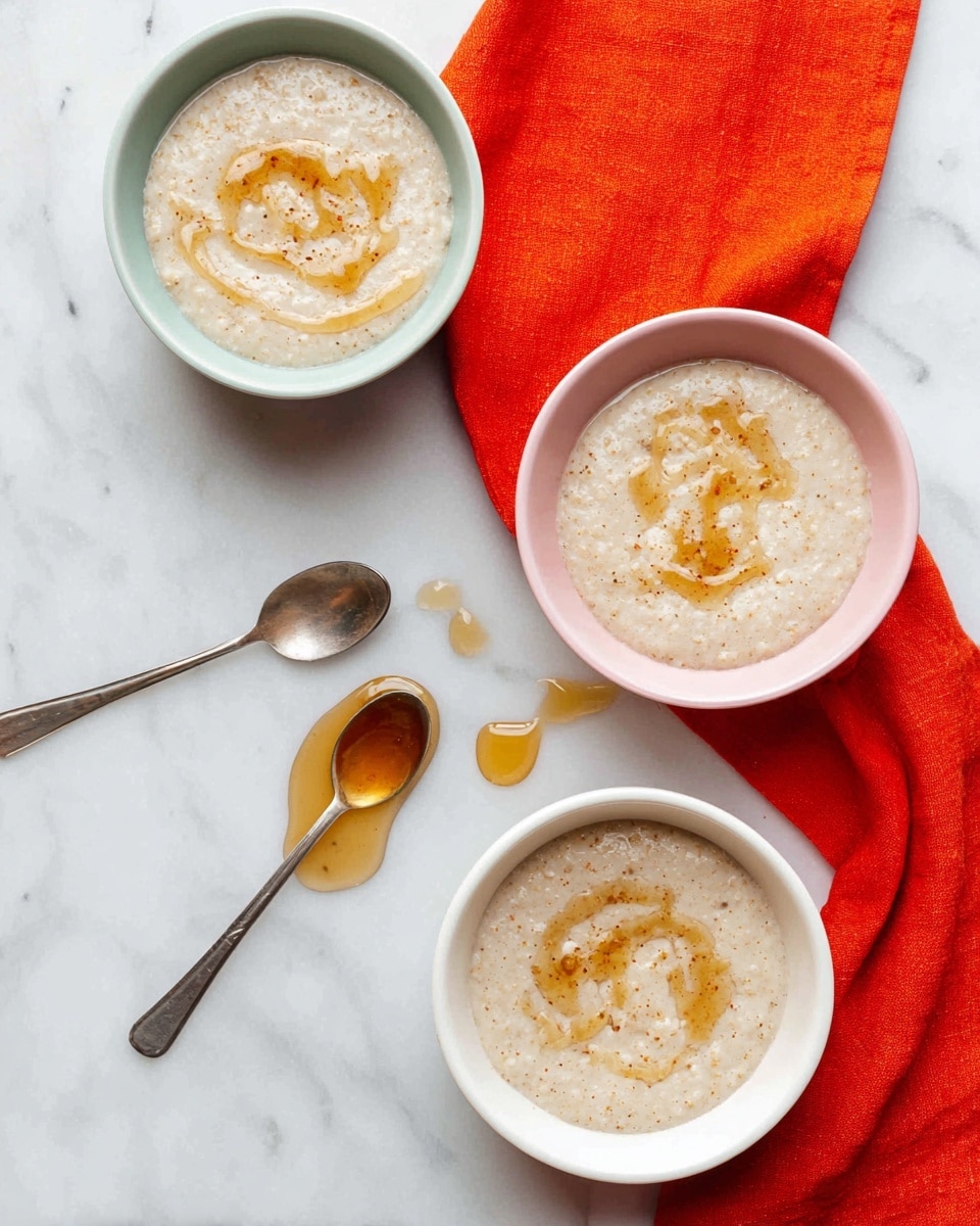 Three bowls filled with a creamy, beige porridge that has a swirl of honey on top. The bowls sit on a white marbled surface. Two of the bowls are white, and one is a light pink color. Under one white bowl is a bright red-orange cloth. A silver spoon with some honey on it lies between the bowls, with a few small drops of honey spilled on the surface nearby. The porridge looks smooth with small grains visible in the texture. photo taken with an iphone --ar 4:5 --v 7