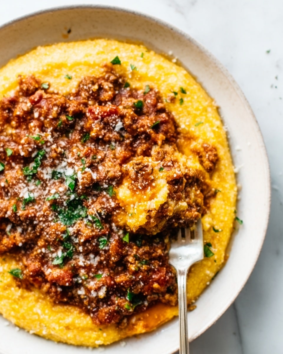 The image shows a round white plate with a base layer of smooth, golden yellow polenta that looks creamy and soft. On top of the polenta is a thick layer of rich, brown red meat sauce speckled with small bits and herbs, giving it a chunky texture. A silver fork is partially stuck into the dish on the right side, resting on the polenta and sauce. The dish is sprinkled with grated white cheese and drops of chopped green herbs for color contrast. The background is a white marbled surface. Photo taken with an iphone --ar 4:5 --v 7