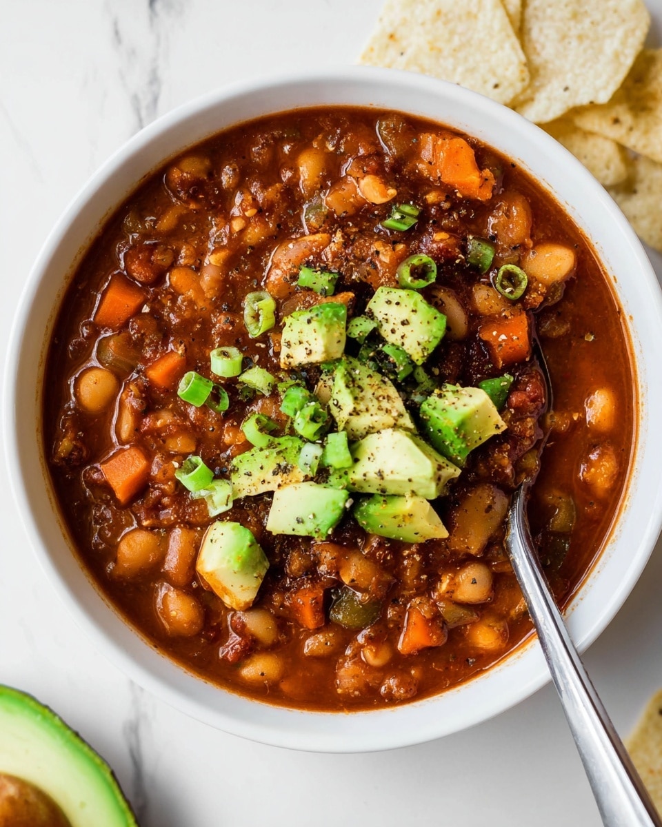 A white bowl filled with rich brown soup containing visible beans and diced tomatoes as the first layer, topped with a layer of shredded orange-yellow cheese that covers the center. Above the cheese, there are chunks of green avocado pieces arranged neatly on one side, with small chopped green onions sprinkled across the top. Scattered broken pieces of light beige tortilla chips are spread over the cheese, giving a crunchy texture. The bowl sits on a white marbled surface. In the background, another similar bowl of the same soup is slightly out of focus. Photo taken with an iphone --ar 4:5 --v 7