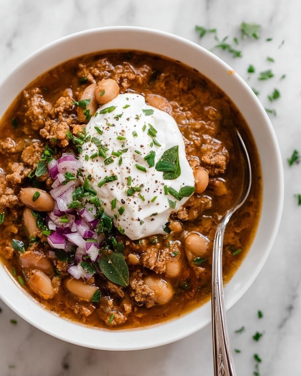 A white bowl filled with a thick, brown stew made of crumbled meat and white beans in a rich sauce, topped with a large dollop of white sour cream sprinkled with chopped green herbs and black pepper, scattered with small pieces of chopped purple onion and fresh green chives. A silver spoon is partially submerged in the stew, resting against the side of the bowl. The bowl is placed on a white marbled surface with small green herb pieces around it. Photo taken with an iphone --ar 4:5 --v 7