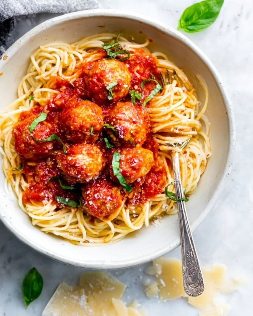 The image shows a white bowl filled with long, cooked pasta noodles arranged in a circle. On top of the noodles are six round meatballs covered in a thick, bright red tomato sauce, sprinkled lightly with grated cheese. There are small green basil leaves around the meatballs adding a pop of color. A silver fork rests on the side of the bowl, partially inside the noodles. The bowl sits on a white marbled surface. Photo taken with an iphone --ar 4:5 --v 7