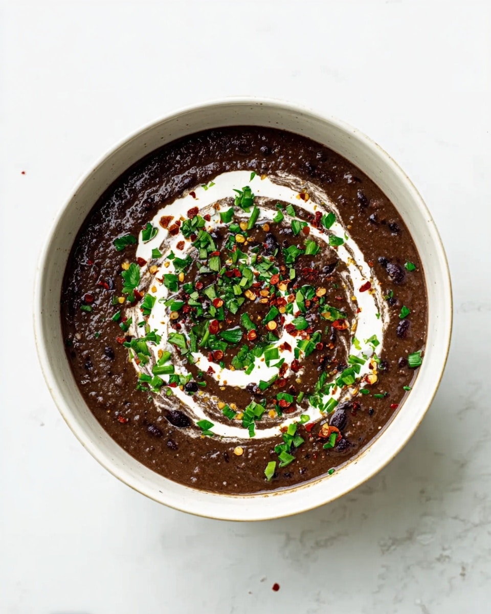 A white bowl filled with dark brown black bean soup that has a thick and textured look. On top, there is a swirl of white cream spread across the center in a rough circle. Green chopped herbs and small red pepper flakes are scattered over the cream and soup, adding color and freshness. The bowl sits on a white marbled surface, showing some light shadows around it. Photo taken with an iphone --ar 4:5 --v 7