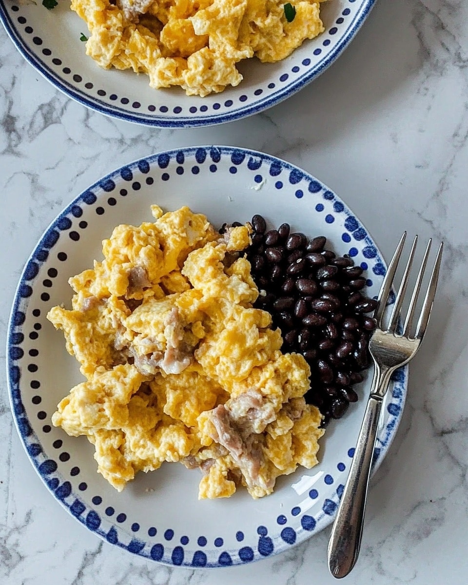 The image shows two white plates with a blue dotted edge set on a white marbled surface. Each plate holds scrambled eggs mixed with pieces of a light brown cooked meat scattered evenly within the eggs, giving a textured, fluffy appearance with a slightly yellow and golden color. Next to the eggs on the plate in the foreground, there is a heap of small, shiny black beans, contrasting with the eggs' lighter colors. A silver fork rests on the right side on the plate with black beans, positioned just above the eggs. The plate in the background holds just the scrambled egg and meat mix. The lighting highlights the soft texture and mild shine of the eggs and beans. The photo taken with an iphone --ar 4:5 --v 6.1