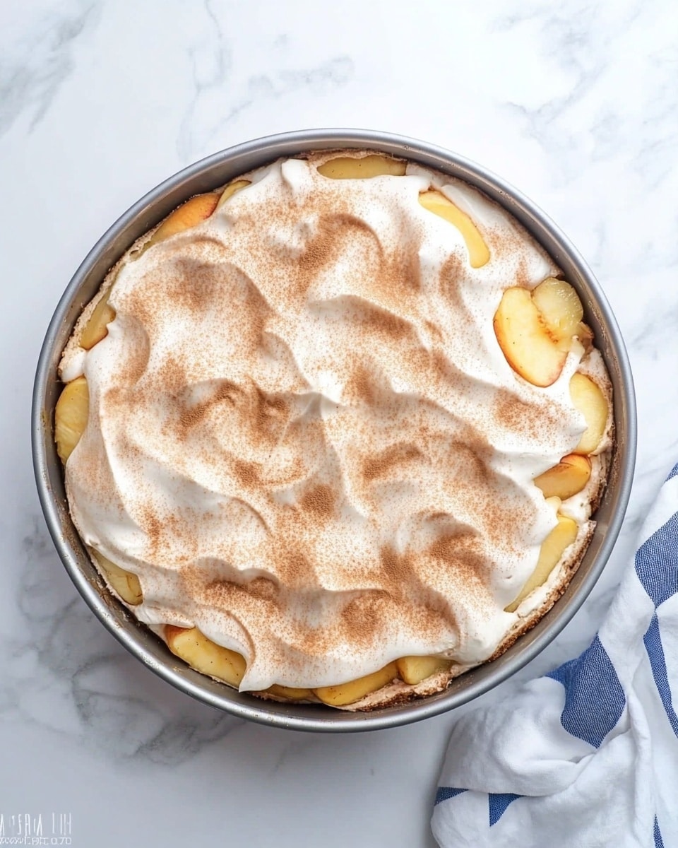 A round metal baking pan filled with a layered dessert placed on a white marbled surface, the bottom layer has small yellow-orange fruit pieces, likely peaches, visible around the edges. On top is a thick layer of light, fluffy cream or meringue spread unevenly with soft swirls and dusted with a light brown powder, possibly cinnamon. A white and blue checkered cloth is partially visible on the right side of the image. Photo taken with an iphone --ar 4:5 --v 6.1