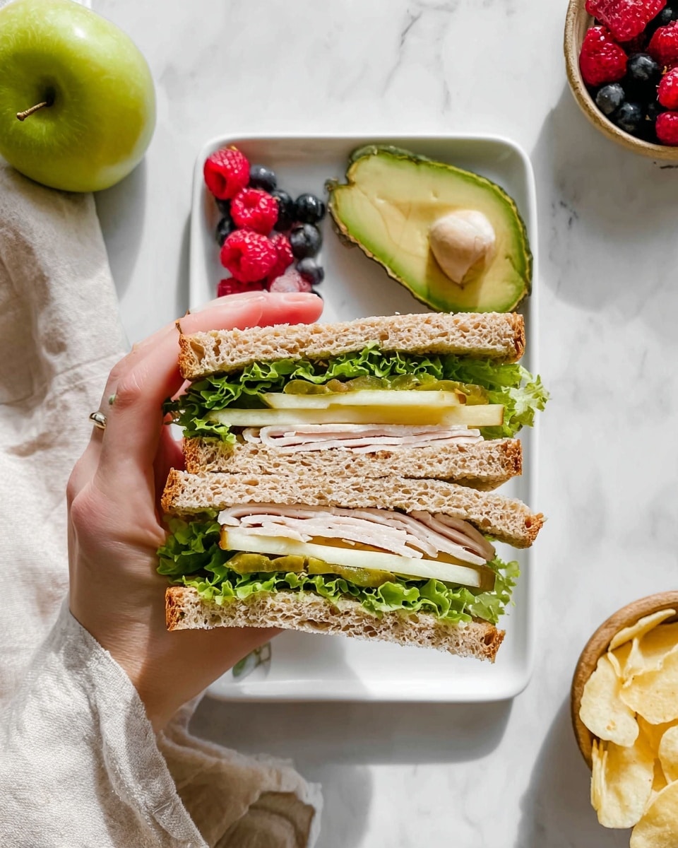 The image shows a sandwich held by a woman's hand over a white rectangular plate on a white marbled surface. The sandwich has two thick layers of whole grain bread with four visible layers inside: the bottom layer is green lettuce with a leafy texture, followed by thinly sliced pale turkey meat, then slices of pale yellow apple, and topped with more green lettuce and some light green pickles. Around the plate, there is half an avocado, a small bowl with a mix of red raspberries, bright red strawberry, and dark blueberries, a white cloth napkin, a green apple partly sliced, and a small bowl of light yellow potato chips. The photo taken with an iphone --ar 4:5 --v 7