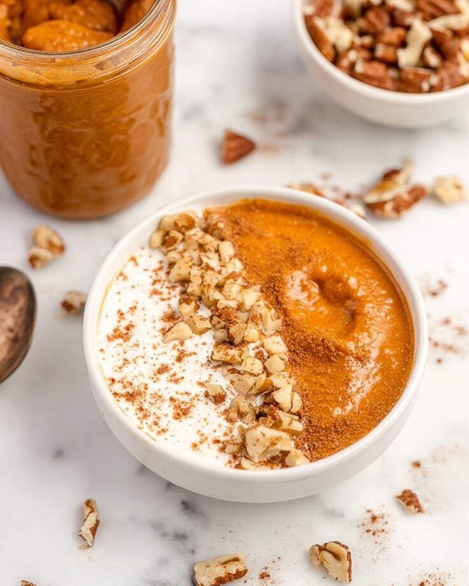 The image shows a round white bowl filled with two distinct layers on top: one side has a smooth, thick orange-brown layer, and the other side has a creamy white layer sprinkled lightly with a brown powder. The orange-brown layer is decorated with chopped light brown nuts along the edge where it meets the white layer. There is a jar in the background that holds more of the orange-brown mixture, and scattered pieces of light brown nuts are spread around the bowl on a white marbled surface. Another white bowl with whole nuts is partially visible in the background. photo taken with an iphone --ar 4:5 --v 7