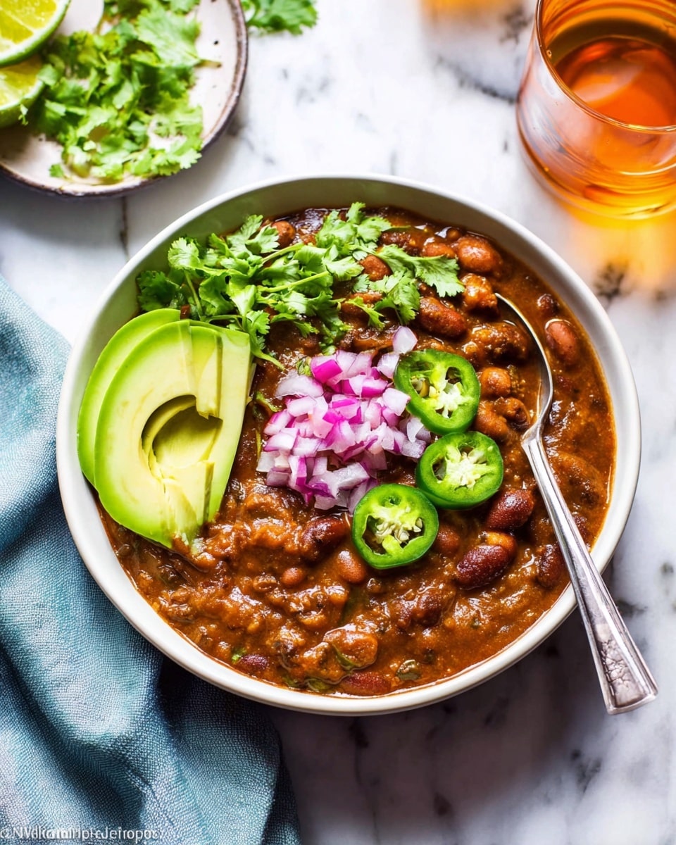 A white bowl holds three main layers: the bottom layer is a thick, brown chili with visible beans and chunks of vegetables, covering nearly the entire bowl. On top of the chili, there's a small pile of shiny, diced red onions and a few thin slices of bright green jalapeño peppers arranged near the center. To the left side of the bowl, there are three thin slices of pale green avocado, fanned out neatly, with fresh green cilantro sprigs resting next to the avocado. A metal spoon is placed inside the bowl on the right side, partially submerged in the chili. The bowl sits on a white marbled surface, with part of a light blue cloth nearby and glasses with amber liquid around. Photo taken with an iphone --ar 4:5 --v 7