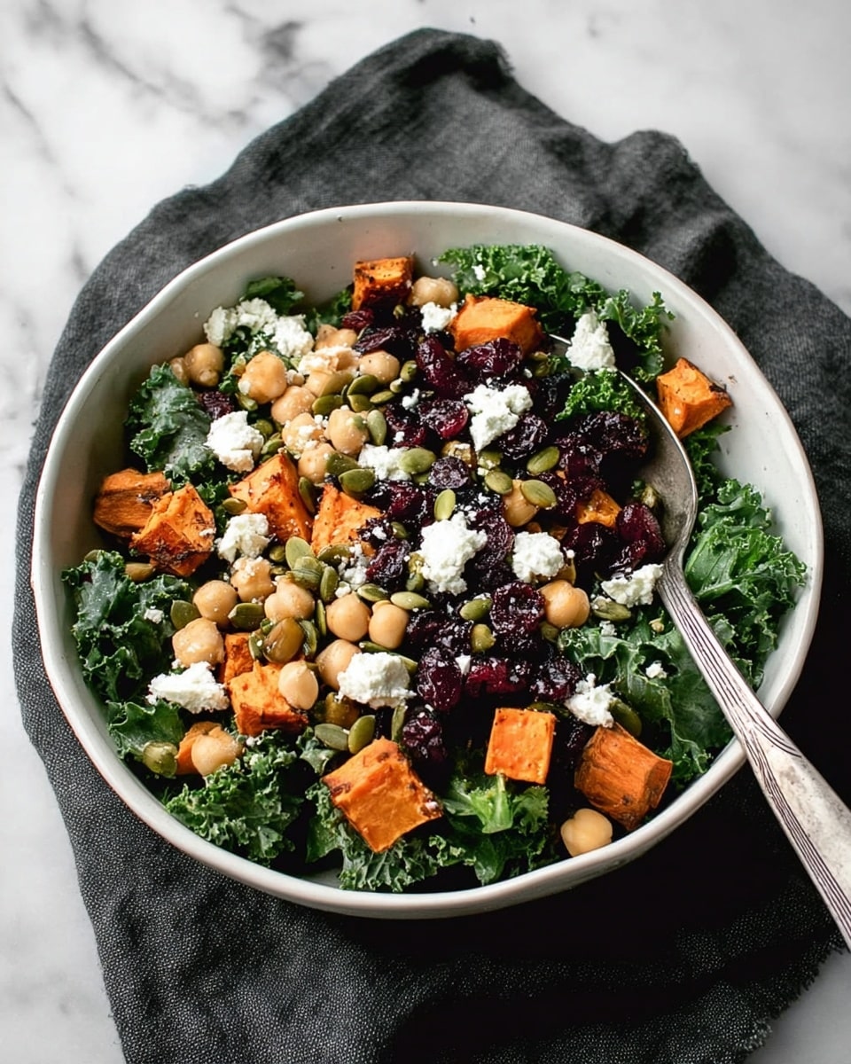A white bowl filled with a colorful salad sits on a white marbled surface with a dark cloth underneath. The salad has three main layers: the bottom layer is dark green curly kale leaves, the middle layer includes light brown chickpeas and bright orange sweet potato cubes, and the top layer has white crumbled cheese, dark dried cranberries, and green pumpkin seeds scattered throughout. A silver spoon rests inside the bowl among the salad ingredients. Photo taken with an iphone --ar 4:5 --v 7