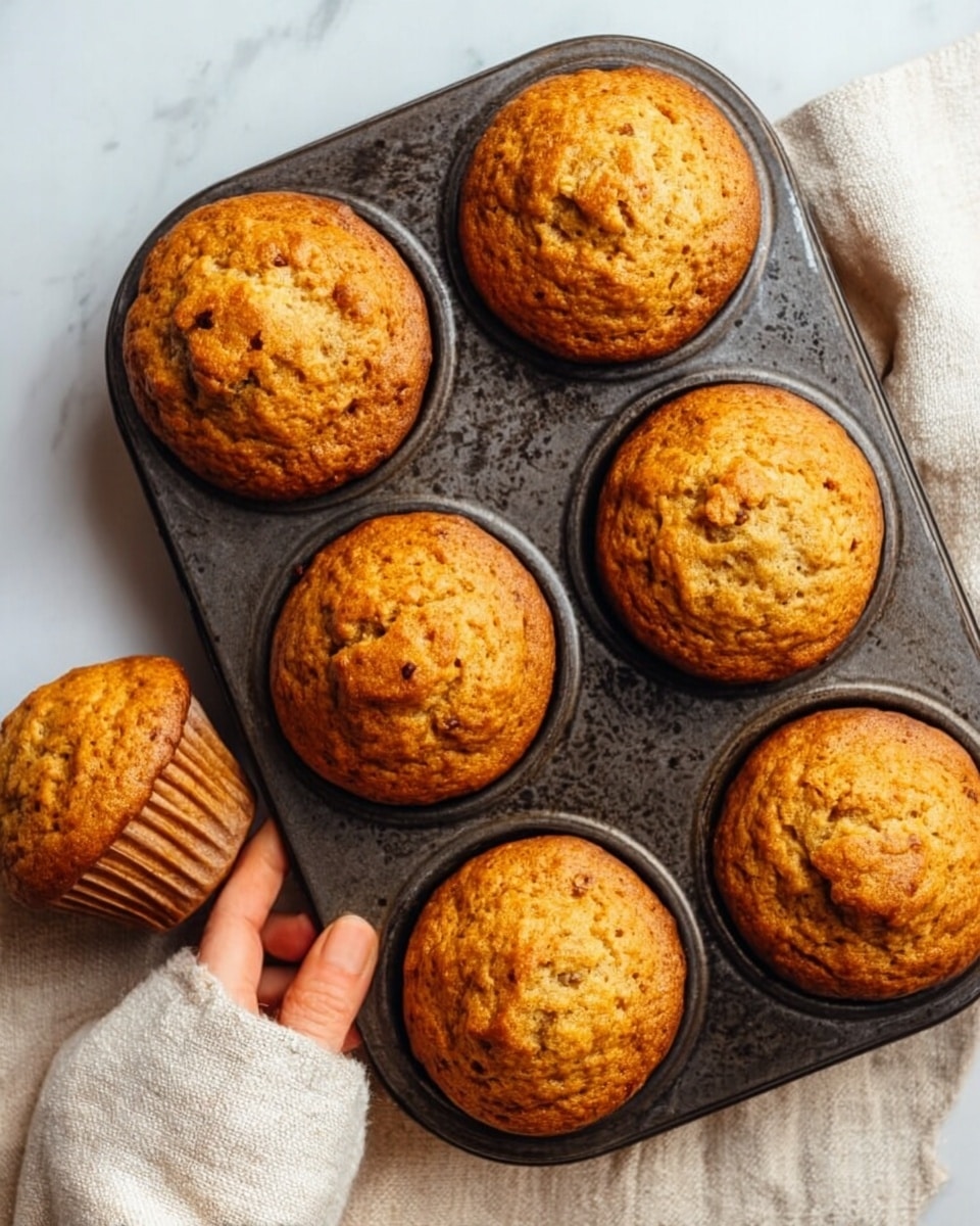 A close-up top view of six brown muffins in a dark metal muffin tray, with one muffin removed and split open in two halves showing a moist, dense texture inside with small bits. The muffins are round and slightly domed with a speckled brown surface. They sit on a light cloth on a white marbled texture background. Next to the muffins are a few crumpled white paper liners. The scene captures a warm, fresh-baked feeling. Photo taken with an iphone --ar 4:5 --v 7