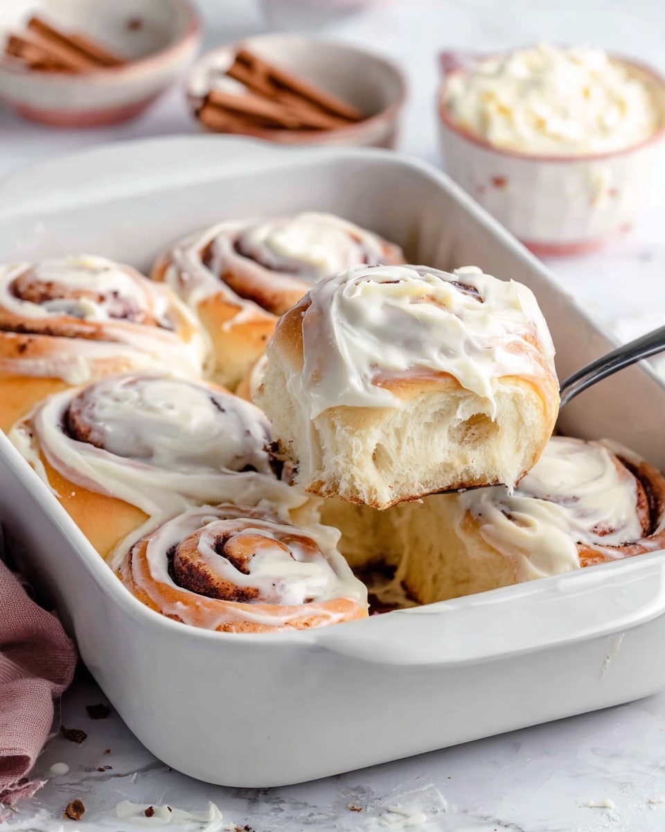 The image shows a white baking dish holding seven soft cinnamon rolls covered generously with creamy white frosting. The rolls are golden brown on the bottom and lightly browned on top, with spiral layers of dough and cinnamon visible. The frosting is thick and creamy, spread unevenly over each roll, melting slightly into the swirls. A silver spoon lifts one roll from the dish, showing its soft, fluffy inside with a light beige color. The background has a white marbled texture with small bowls holding cinnamon sticks and extra frosting slightly blurred in the distance. photo taken with an iphone --ar 4:5 --v 7
