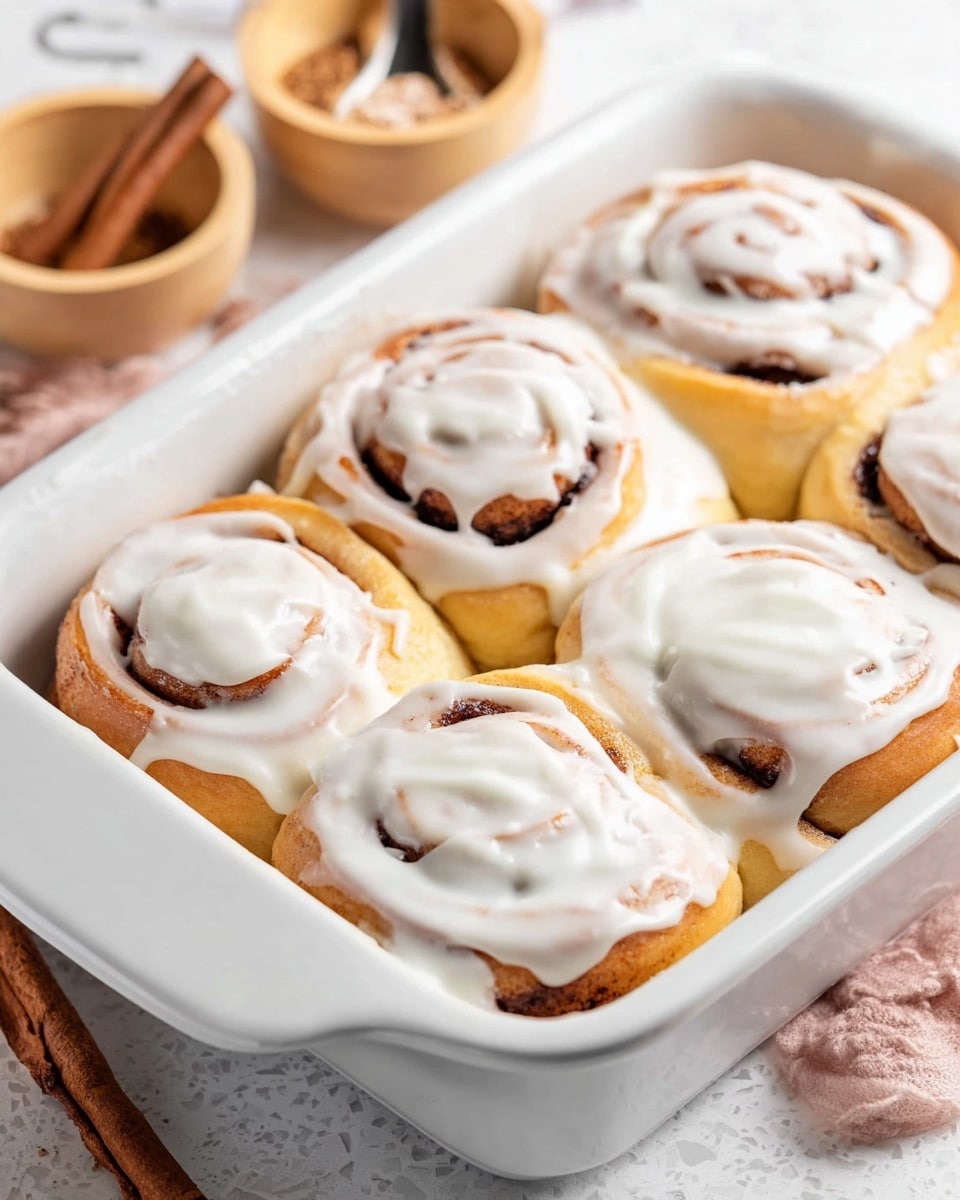 A soft, thick cinnamon roll sits on a white plate with a white marbled surface underneath. The roll has three visible layers: the bottom layer is golden brown baked dough with a fluffy, airy texture; the middle layer is a swirl of dark brown cinnamon filling; the top layer is a thick, creamy white icing spread all over the roll, slightly melting and glossy, covering the cinnamon swirl and edges. Another similar roll on a white plate and a white baking dish with more rolls are blurred in the background. Photo taken with an iphone --ar 4:5 --v 7