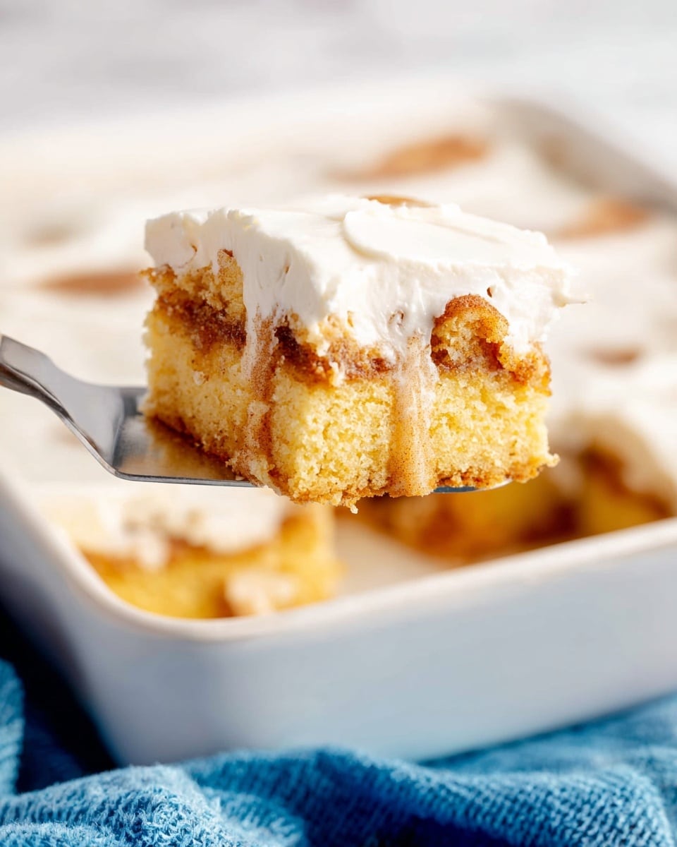 A square piece of layered cinnamon cake is shown on a silver spatula, with a white creamy frosting layer on top that looks smooth and thick. Below the frosting, two layers of soft yellow cake are mixed with a light brown cinnamon swirl that adds texture and color contrast within the cake. The cake is held above a white baking dish filled with more pieces of the same cake, set on a white marbled surface with a blue cloth nearby. The lighting is bright, showing the moist and fluffy texture of the cake layers clearly. Photo taken with an iphone --ar 4:5 --v 7