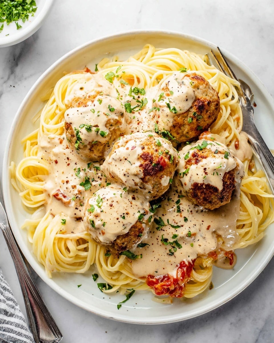 A round white plate on a white marbled surface holds a serving of long pale yellow spaghetti arranged in a loose pile as the base layer. On top, six browned meatballs, each covered with a creamy light beige sauce with small green herb pieces, form the next layer. Small bits of red tomato sauce peek from underneath the sauce and meatballs, adding color contrast. The meatballs are sprinkled with finely chopped fresh green herbs and a dusting of black pepper, adding texture and detail. A fork and a knife rest next to the plate on the surface. photo taken with an iphone --ar 4:5 --v 7