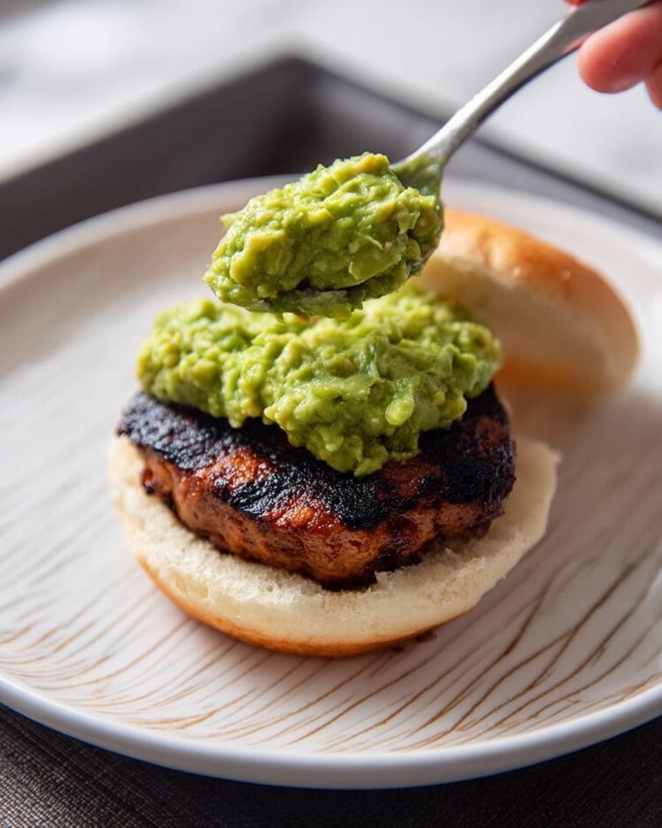 Three small sandwiches sit on a long white plate with a dark background behind. Each sandwich has a soft light brown bun. The bottom layer inside is green mashed avocado spread, thick and fluffy. On top of the avocado is a layer of dark grilled meat with a textured surface. The left and right sandwiches each have two green olives with pimento on top of the bun, stuck by toothpicks. The middle sandwich has two toothpicks with one green olive each, inserted into the top bun, standing upright. Behind the plate is a packet of grilled chicken thighs with an orange, green, and black design. The surface the plate sits on is a white marbled texture. photo taken with an iphone --ar 4:5 --v 7
