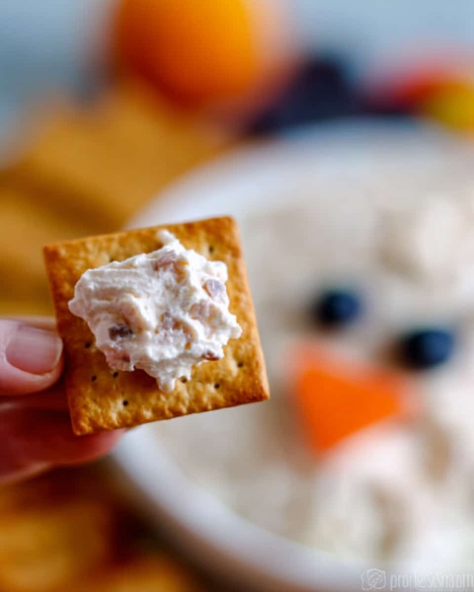 A close-up of a single square golden-brown cracker held by a woman's hand, topped with a dollop of creamy white spread with small darker bits visible inside it. In the background, there is a blurred look of a white bowl with a dip that has a face made from colorful ingredients; soft orange shapes form the eyebrows, two dark blue round items create the eyes, and a small orange piece acts as the nose, all sitting on a creamy white dip. The scene is set on a white marbled surface. photo taken with an iphone --ar 4:5 --v 7