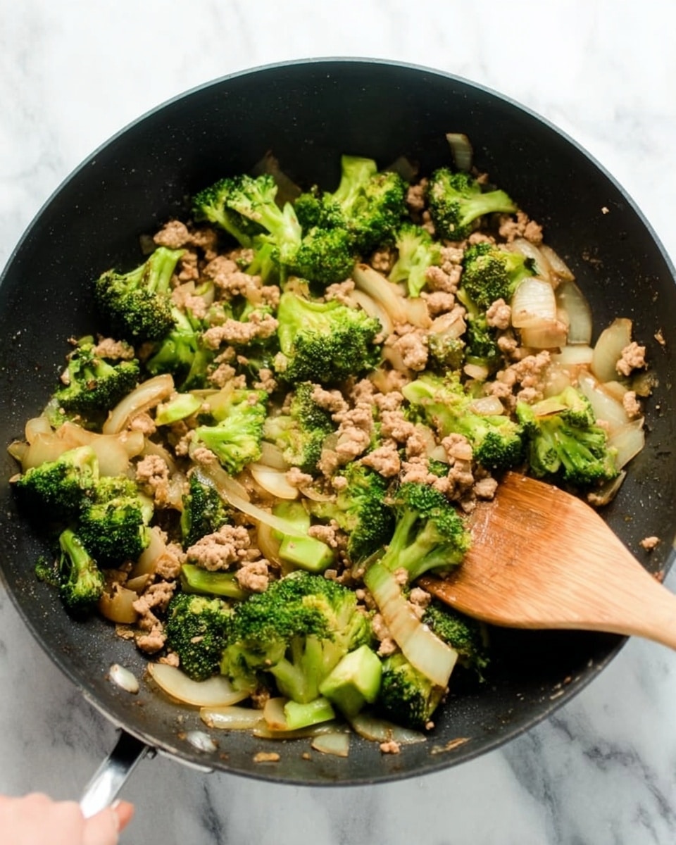 This image shows a white bowl filled halfway with plain white rice on one side, and topped on the other side with a mix of lightly cooked ground meat, broccoli florets, and soft pieces of cooked onion. The broccoli is bright green, and the meat is light brown, with the translucent onion slices scattered throughout. A silver spoon is placed inside the bowl, with its handle resting on the edge. The bowl sits on a white marbled surface, and part of another white bowl full of the same dish is slightly visible in the background. photo taken with an iphone --ar 4:5 --v 7