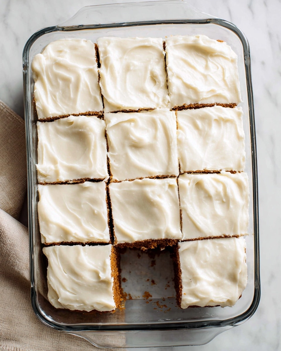 The image shows a rectangular clear glass baking dish filled with a brown cake base topped with a thick layer of smooth white frosting. The cake is cut into nine square pieces with one piece missing from the bottom right corner, revealing the dense brown cake underneath. The frosting is evenly spread with soft swirled textures on each piece. The dish sits on a white marbled surface with a beige cloth visible at the bottom left edge. photo taken with an iphone --ar 4:5 --v 7