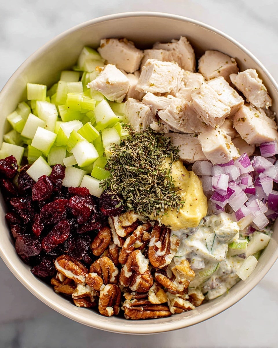 In a white bowl, there are many layers of ingredients placed next to each other in sections. Starting from the top, light beige pieces of cooked chicken sit next to small green diced celery pieces. On the right side, there are small light green apple cubes. Below them, finely chopped red onion pieces create a purple layer next to a dark red heap of dried cranberries. Below that, there is a layer of chopped pecans with a rough texture and brown color. In the middle, there is a creamy yellow dollop of mustard next to a white sauce that looks creamy. On top of the chicken and some other ingredients, there are dark green dried herbs sprinkled evenly. The bowl sits on a white marbled surface photo taken with an iphone --ar 4:5 --v 7