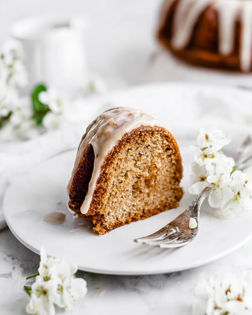 A single slice of brown bundt cake with a light layer of white glaze on top sits on a white round plate. The cake has two visible layers with a slightly darker middle band, showing its texture. A silver fork rests next to the cake on the plate. The background is a white marbled texture, with soft white flowers placed around the plate. Photo taken with an iphone --ar 4:5 --v 7