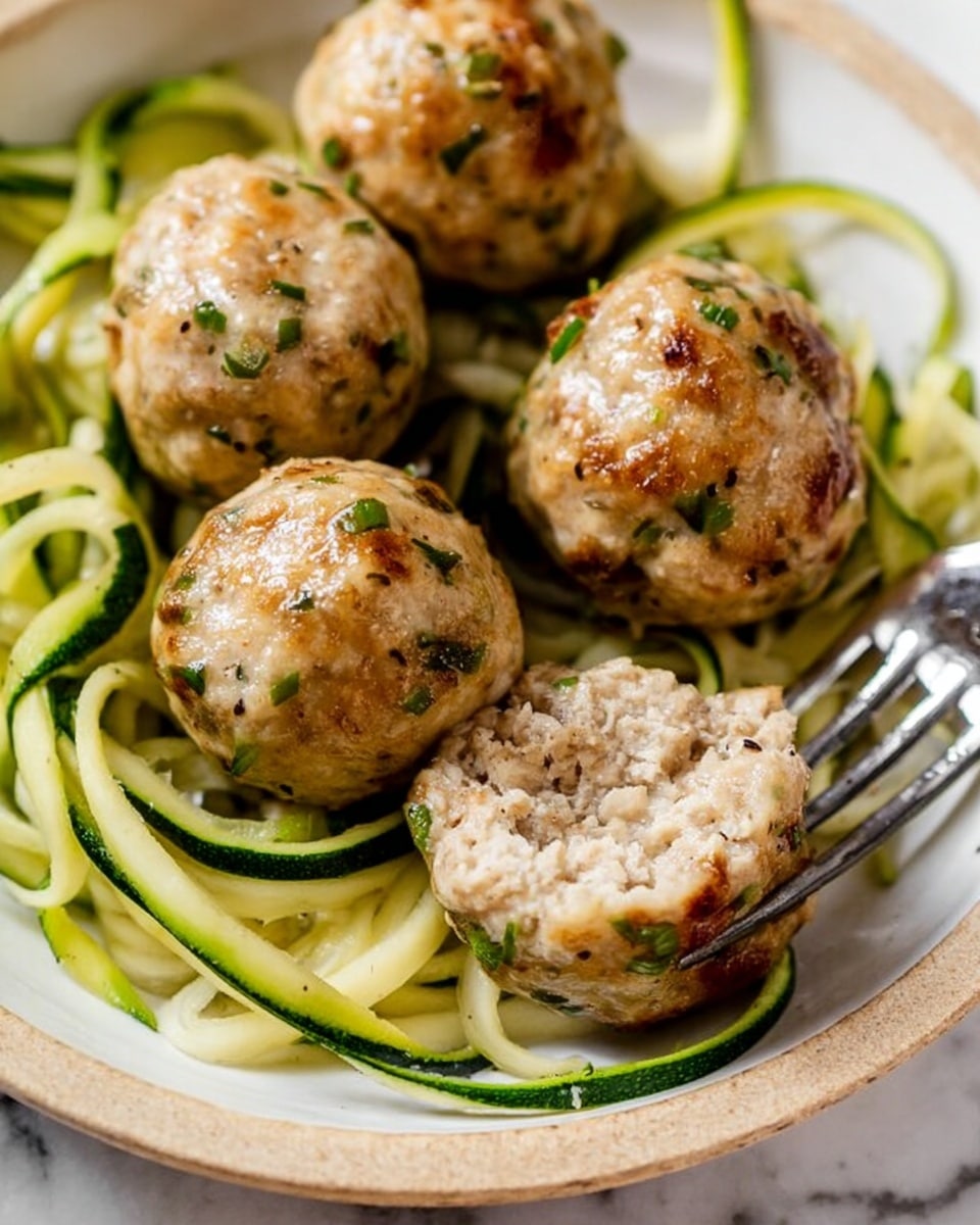 The image shows five round meatballs, light brown with small green herb flecks, sitting on a white plate. One meatball is cut in half, revealing a soft, moist inside. Underneath the meatballs are green spiral vegetables that look like zucchini noodles, curling loosely across the plate. A silver fork is placed near the cut meatball, slightly piercing the noodles. The background is a white marbled surface. photo taken with an iphone --ar 4:5 --v 7