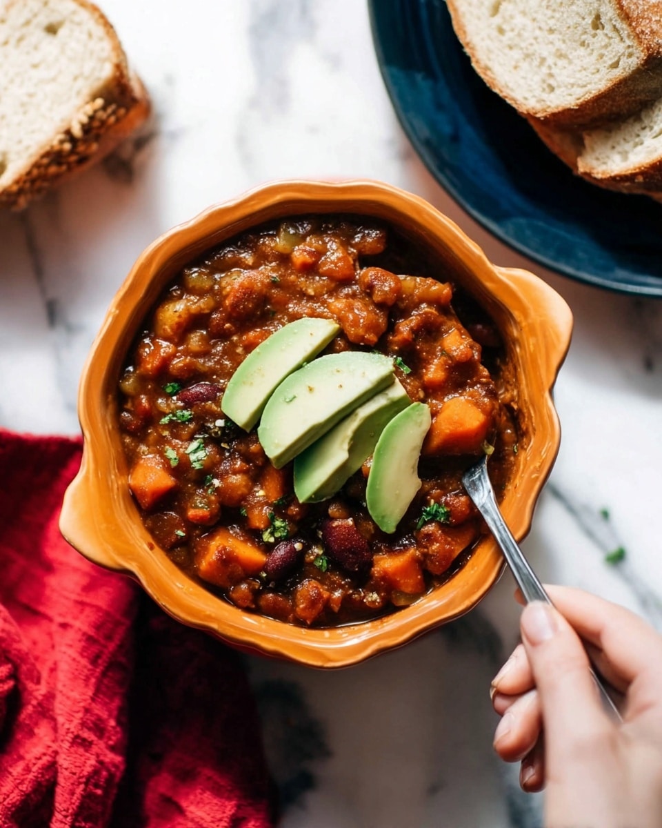 A scalloped orange bowl filled with chunky vegetable chili showing a thick, brownish-red sauce mixed with visible pieces of beans, diced carrots, and other veggies. On top of the chili are three fresh green avocado slices, placed in the center. A silver spoon is partially dipped into the chili, held by a woman's hand on the right side of the bowl. The bowl sits on a white marbled surface, with a dark blue plate holding thick slices of bread positioned in the upper right corner. In the lower left corner, there is a slice of bread partly visible. There is also a red cloth in the top left of the image. The lighting is bright, showing the rich textures of the chili ingredients clearly. Photo taken with an iphone --ar 4:5 --v 7