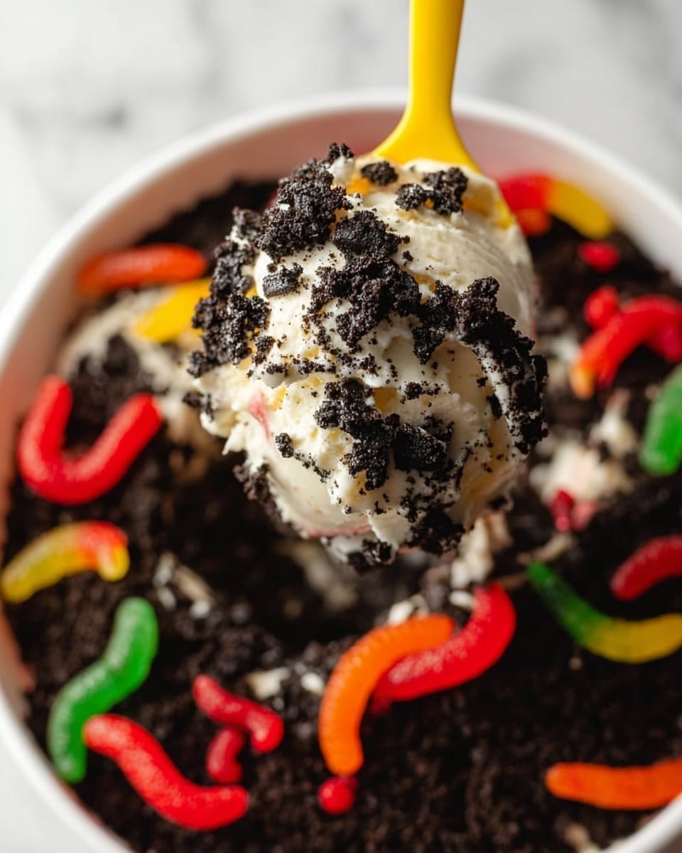 A close-up of a yellow spoon holding a scoop of creamy white ice cream covered with crushed black cookie crumbs. The scoop is above a white bowl filled with more crushed black cookie crumbs and colorful gummy worms in red, orange, and green, scattered around to look like worms in soil. The background is a white marbled surface. Photo taken with an iphone --ar 4:5 --v 7